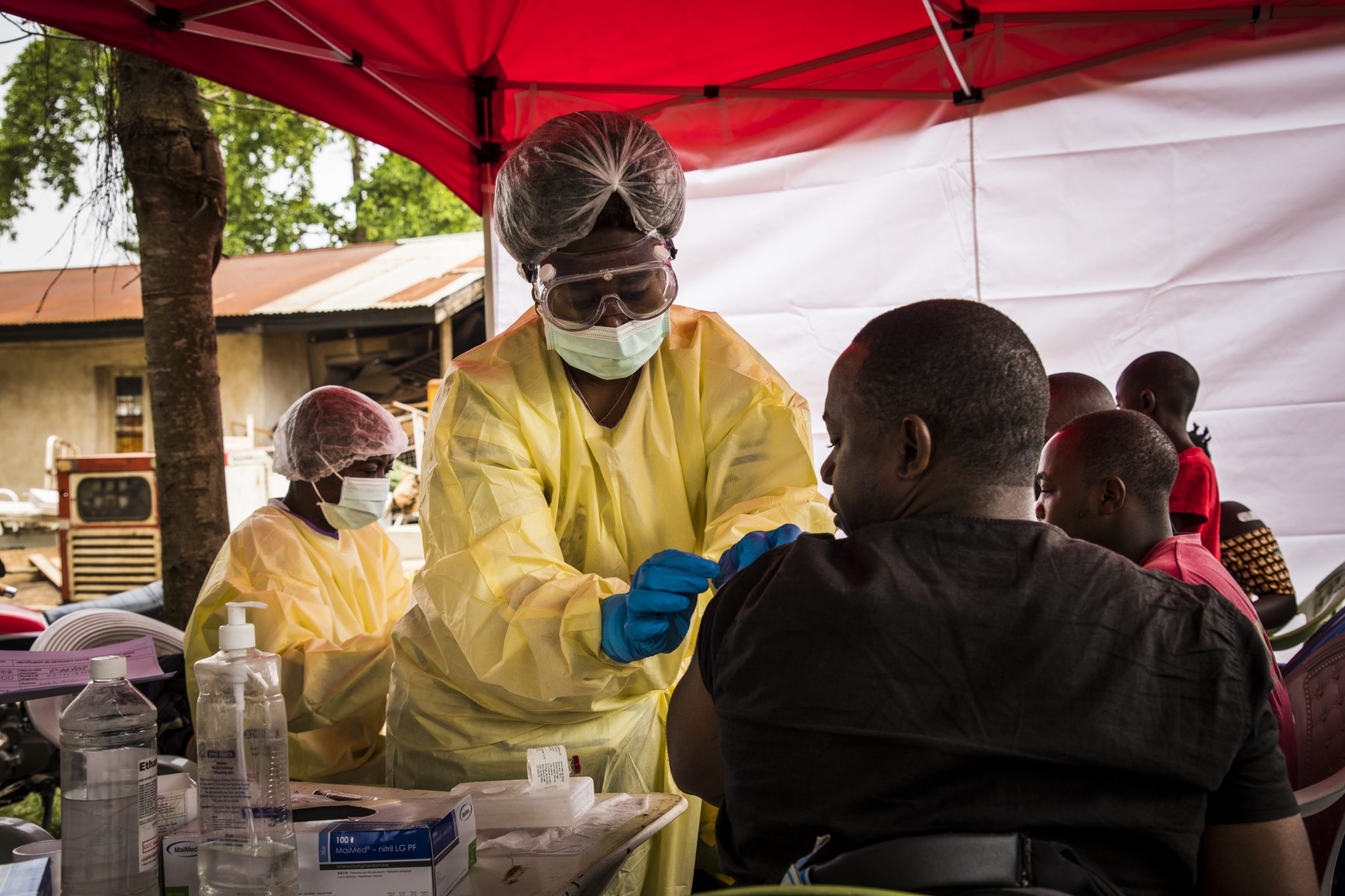 A nurse dressed in safety clothing and with a mask administers an injection to a man